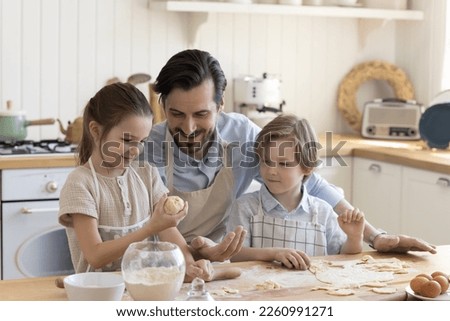 Happy positive dad and little son and daughter kids in aprons baking in kitchen, shaping dough at floury kitchen table, preparing bakery food, dessert, cooking together, talking, smiling Royalty-Free Stock Photo #2260991271