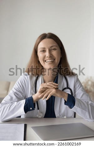 Happy beautiful practitioner young woman posing in office, sitting at desk, looking at camera with toothy smile. Positive successful doctor with stethoscope head shot portrait Royalty-Free Stock Photo #2261002169