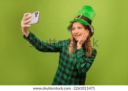 Happy smiling girl in green leprechaun hat and checkered plaid shirt taking selfie photo on smartphone or mobile phone and holding hand on face or cheek on green background.

St Patricks day concept.