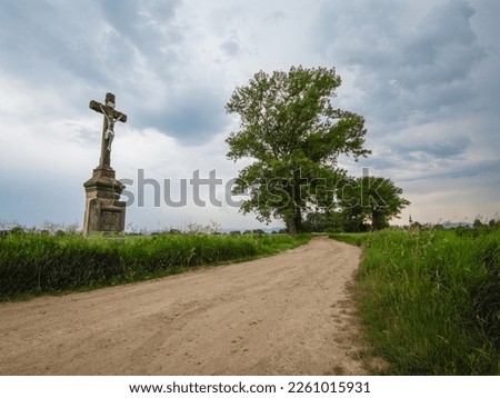 Field, dirt road with a roadside cross. Summer, rural landscape with stone cross, meadow and trees. Cloudy weather, no body, Poland.