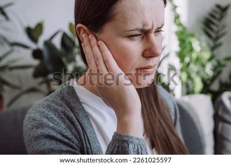 Closeup up side profile of sick young caucasian female having ear pain touching painful head sitting on sofa. Unhealthy woman 20s suffering from painful otitis. Health care and tinnitus concept Royalty-Free Stock Photo #2261025597