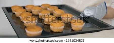 Hand of baker wearing mitten, holding tray with muffins. Fresh baked cupcakes, bakery food for birthday party, dessert, breakfast, cakes arranged in rows on metal pan. Close up banner shot