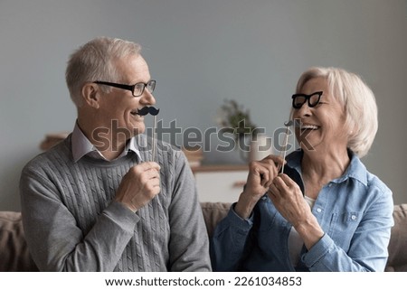 Happy joyful older couple of grandparents playing with photo shooting props, applying fake moustache and glasses on sticks to faces, having fun, smiling, laughing