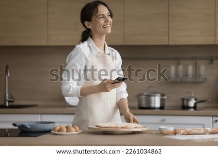 Cheerful bakery blogger woman holding smartphone at table with raw pastry ingredients, preparing pizza in home kitchen, looking away with dreamy smile, thinking, laughing