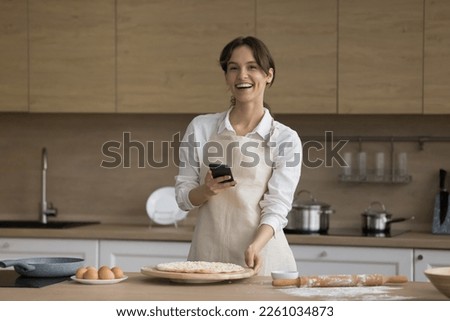 Happy cook blogger girl holding mobile phone over raw bakery food on floury kitchen table, looking at camera, smiling, laughing. Cheerful young baker woman taking picture of food on cell