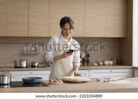Happy young cook girl wearing apron, using mobile phone in home kitchen while baking pie, pizza, pastry, taking photo of raw bakery food for blog, chatting, making call