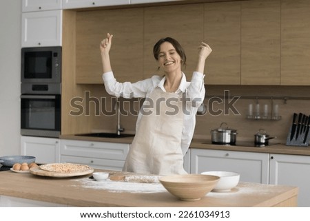 Happy active inspired baker girl in apron dancing to music at floury table with bakery, pizza ingredients, dough, eggs, dish, smiling, laughing, enjoying cook job, chef profession Royalty-Free Stock Photo #2261034913