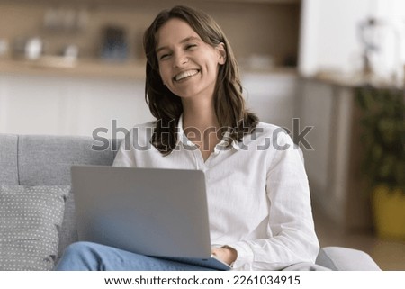 Cheerful beautiful laptop user woman sitting on home couch, holding computer on lap, typing, looking away, smiling, laughing, enjoying wireless Internet connection technology