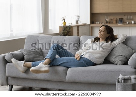 Positive peaceful young woman resting on sofa at home, leaning on back, breathing fresh air, looking at window away, enjoying break, leisure, thinking, dreaming. Full length shot Royalty-Free Stock Photo #2261034999