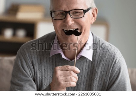 Happy positive senior man applying fake paper moustache on stick to face, celebrating movember, looking at camera, having fun, smiling, laughing, using photo shooting prop. Cropped portrait Royalty-Free Stock Photo #2261035081