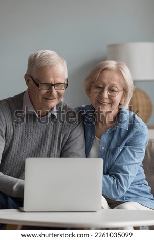 Happy elderly husband and wife in glasses using online app on laptop, typing, talking on video call, smiling, laughing, shopping on internet, sitting on home sofa, enjoying Internet communication Royalty-Free Stock Photo #2261035099