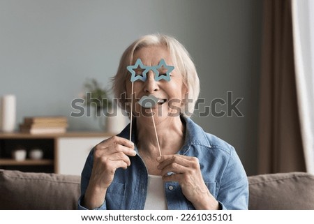 Cheerful happy senior woman applying fake paper moustache and goggles on stick to face, looking at camera, having fun, smiling, laughing, using prop for photo shooting