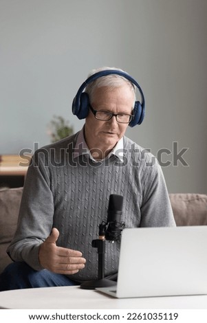 Serious mature influencer recording speech for blog, broadcasting from home, giving online workshop. Older man in wireless headphones, speaking at professional mic and laptop. Vertical shot