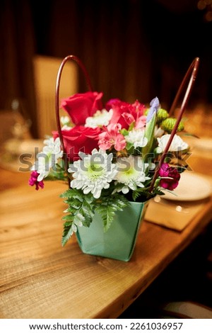 a decorative paper basket with bright flowers stands on the table. A basket, a paper bag with bright flowers stands on a table in a cafe, restaurant. gift, wedding, birthday