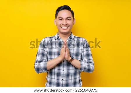 Young handsome Asian man wearing a plaid shirt gives greeting hands with a big smile isolated on yellow background. People lifestyle concept