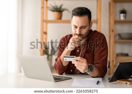 Serious business man sitting at table in home office, holding and looking at credit card, worried about debt and credit card deficit, considering a financial loan from bank. Royalty-Free Stock Photo #2261058275