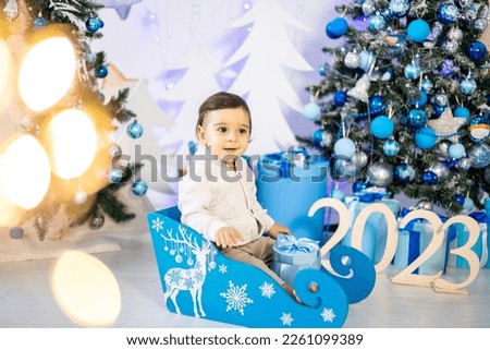 a small child, a boy in a knitted sweater, is sitting in a sled against the background of a festive decorated Christmas tree at home, smiling. New Year 2023, Christmas