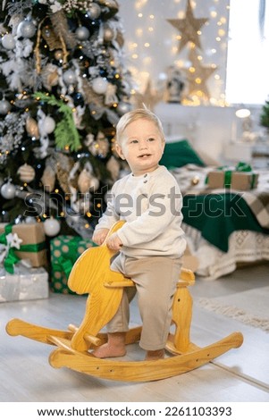 a cute little boy in a Santa costume rides a rocking toy under a festive Christmas tree with gifts in the living room of the house, a happy kid celebrates Christmas and New Year at home