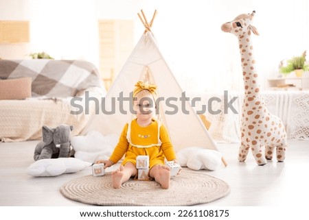 a cute healthy girl is sitting on a rug in the bright living room of the house against the background of a wigwam and plush toys, playing with wooden educational toys, home textiles