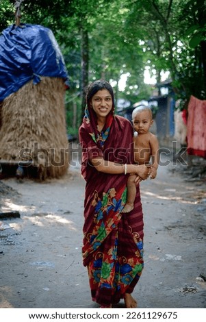 South asian young mother holding her baby son in a village environment, straw hatch in background, woman wearing shari 