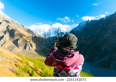 A traveler girl in a hat takes pictures with her back to the camera on the phone of the alpine lake Shavlinskoye with mountains with snow and glaciers in Altai in autumn.
