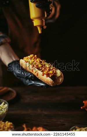 The chef in gloves holds a hot dog. Fast food cooking. Classic hot dog in dark style. Royalty-Free Stock Photo #2261145455