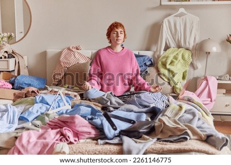 Relaxed ginger young woman meditates on bed breathes deeply sits in lotus pose surrounded by variety of unfitting clothes makes revision of her garment at home collects clothing for donation Royalty-Free Stock Photo #2261146775