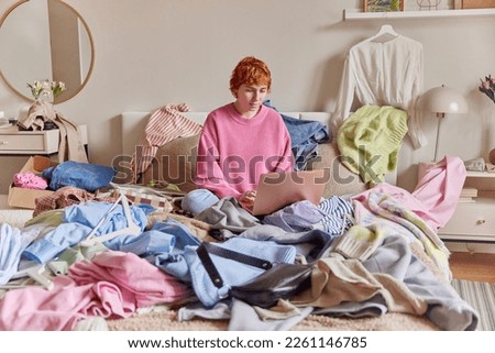 Indoor shot of serious woman with ginger hair wears pink jumper keyboards on laptop computer poses on bed surrounded by variety of clothes sorts out wardrobe. Mess of clothing all over room. Royalty-Free Stock Photo #2261146785