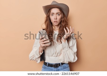 Waist up shot of confused European woman with long wavy hair wears cowboy hat and blouse feels unaware holds mobile phone awaits for call carries bag on shoulder isolated over brown background