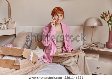 Delivery concept. Serious redhead young woman talks via smartphone holds parcel unpacks cardboard box with bought jumper dressed formally poses on comfortable bed against cozy bedroom interior
