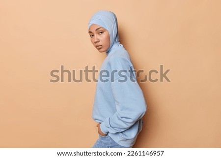 Sideways shot of serious Islamic woman looks directly at camera wears hijab casual pullover and jeans keeps hands in pockets isolated over brown background. Religious female model poses indoor