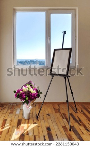 An easel with a picture by the window and flowers in a vase