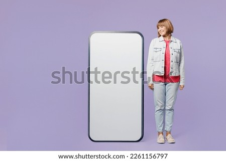 Full body happy elderly blonde woman 50s years old wear casual clothes denim jacket t-shirt look at big huge blank screen mobile cell phone with area isolated on plain pastel light purple background