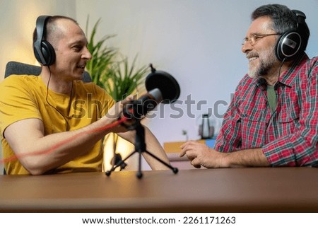 Horizontal view of two radio hosts recording podcast in broadcasting studio. Middle aged Man and mature man podcasters interview each other for radio podcast. headphones laughing while recording 