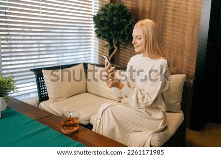 Charming blonde female relaxing on comfortable couch, holding smartphone in hands. Pretty young lady chatting in social networks, watching funny videos, using mobile applications at home.