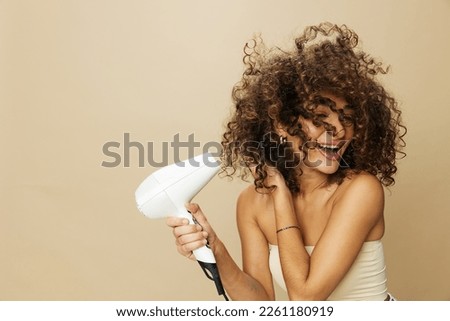 Woman dries curly afro hair with blow dryer, home beauty care styling products hair, smile on beige background Royalty-Free Stock Photo #2261180919