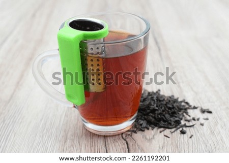 A cup of brewing ceylon tea with tea infusion strainer and pile of tea leaves on wooden table
