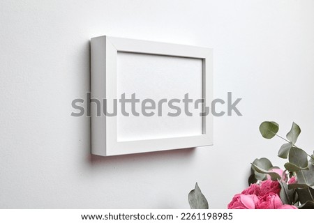 Photo frame mockup hanging on white concrete wall and bouquet of pink roses with eucalyptus leaves. Empty blank picture frame, interior decor