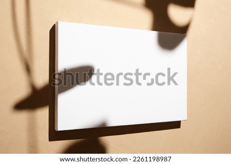 White canvas, blank picture mockup hanging on beige wall with dark shadows of leaves. Poster mock up, empty canvas with shadows of plant, side view