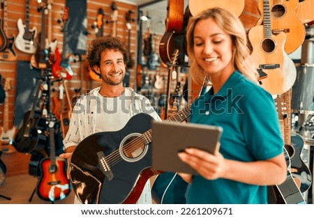 Handsome bearded curly man choosing a guitar in a musical instrument store getting the help of a consultant. Woman seller telling about assortment of guitars to buyer.