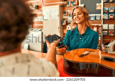 Handsome bearded curly man buying a guitar in a musical instrument store and paying for the purchase using a smartphone. Contactless payment. Woman seller selling a guitar to a buyer.