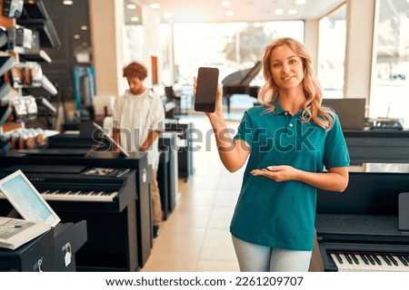 Handsome bearded curly-haired man choosing a piano in a musical instrument store getting the help of a consultant. Woman seller showing screen of smartphone.