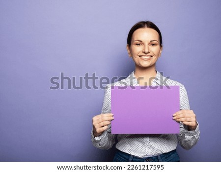 Young brunette woman holding purple blank advertising board standing on lilac background in studio