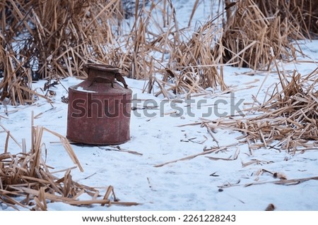 An iron rusty barrel with a lid on the snow among the dry grass, in winter. Industrial canister for use in animal husbandry. Old milk can. Selective focus.