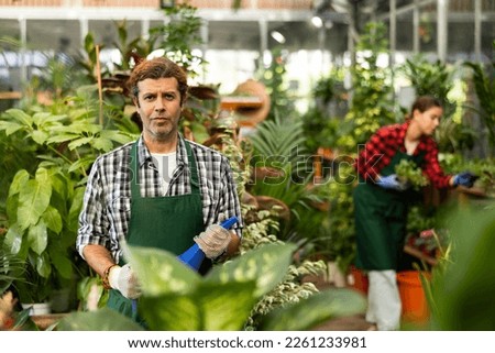 Portrait of positive confident adult floriculturist working in gardening store, standing among greenery of ornamental potted houseplants with watering can in hands.. Royalty-Free Stock Photo #2261233981