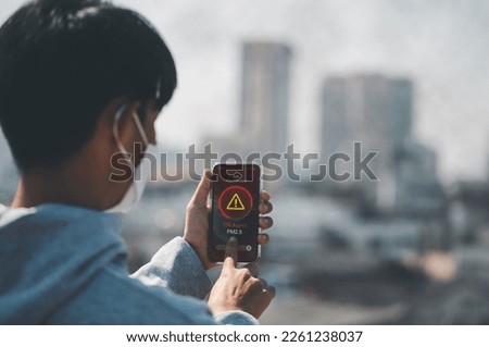 Man wearing the N95 Respiratory Protection Mask and using a smartphone app showing PM 2.5 dust in the capital that exceeds the standard that is harmful to health. air pollution concept, pm 2.5 warning Royalty-Free Stock Photo #2261238037