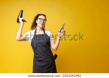 young girl hairdresser in uniform holds a hair dryer and scissors and dances on a yellow background, a woman stylist in an apron shows equipment for work