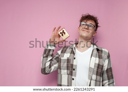 young curly guy with glasses shows a small gift box and dreams on a pink background, a mini gift for a holiday and birthday
