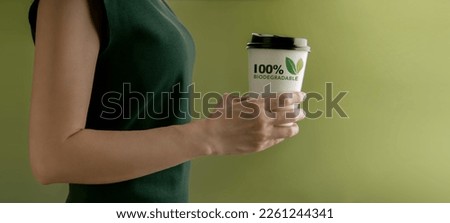 Biodegradable and Compostable Packaging Concept. Closeup of Woman Holding a Hot Cup of Coffee against a Green Wall. Zero Waste Materials. Environment Care, Reuse, Renewable for Sustainable Lifestyle Royalty-Free Stock Photo #2261244341