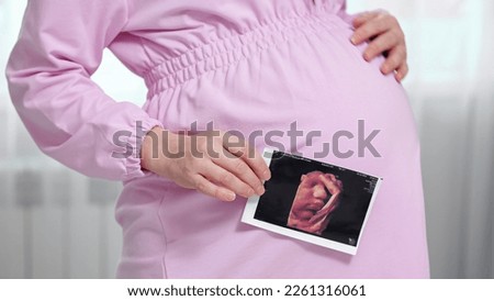 Pregnant woman holds ultrasound photo and awaits for future baby. Gravid lady wearing pink dress enjoys becoming mother and giving birth to baby, side view